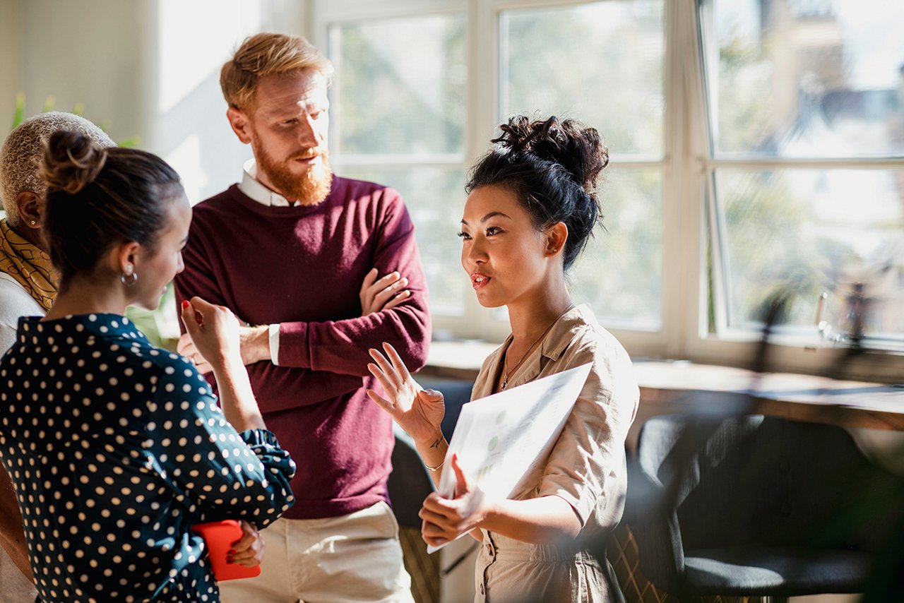 Colleagues standing in a small group discussing something. One of the women is holding documents and gesturing with her hands as the others watch and listen. Colleagues standing in a small group discussing something. One of the women is holding documents and gesturing with her hands as the others watch and listen.