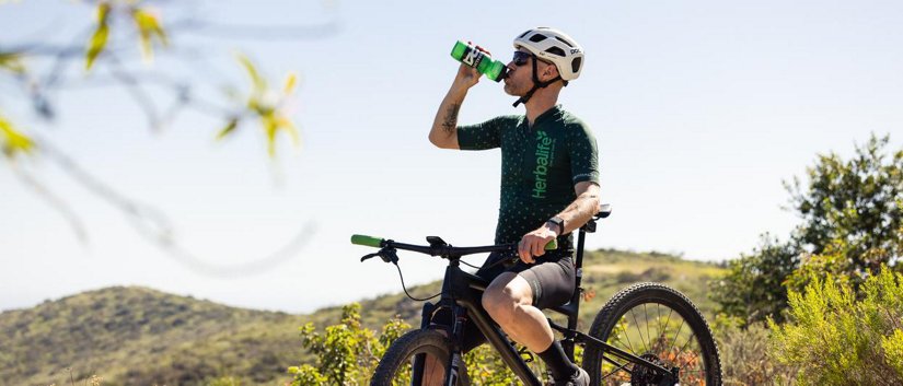 Person wearing helmet drinks from a green bottle with a bike on a hillside; jersey shows Herbalife text.